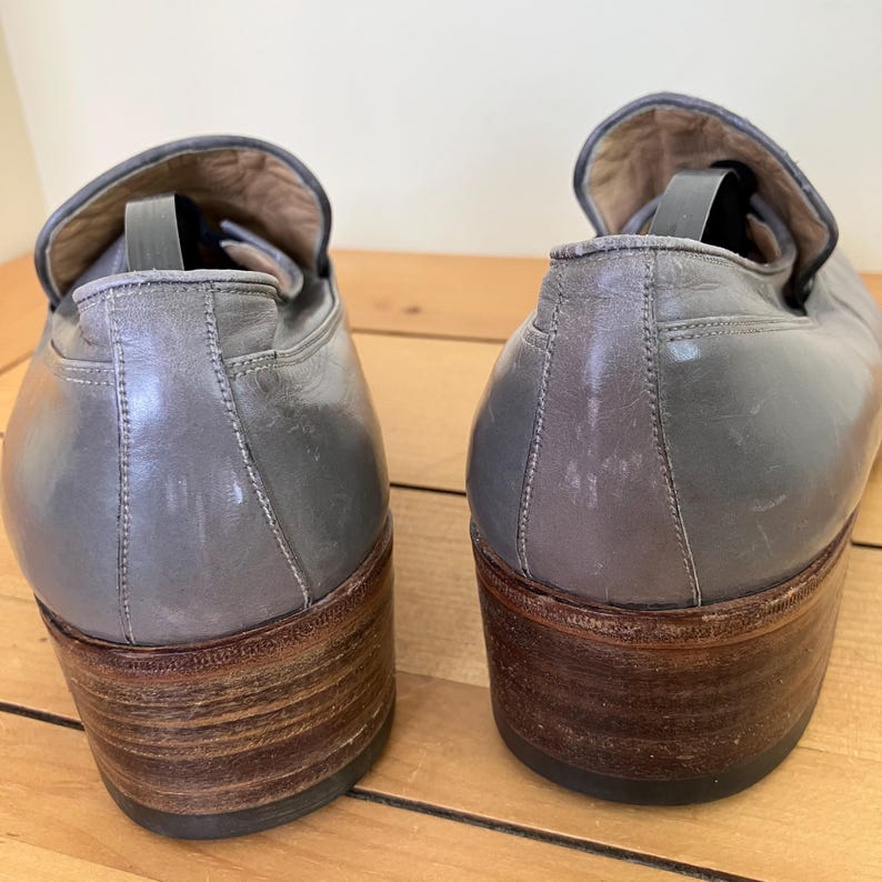 May include: A pair of gray leather loafers with brown wooden heels. The loafers have a simple design and are in good condition.