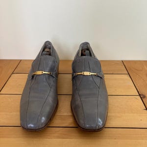 May include: A pair of gray leather loafers with gold buckles. The loafers have a pointed toe and a low heel.