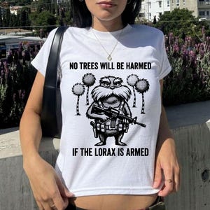 以下が含まれることがあります： 白いTシャツ。黒いグラフィックで、ライフルを持ったロラックスと、トゥーフラの木々が描かれています。テキストは「NO TREES WILL BE HARMED IF THE LORAX IS ARMED」と書かれています。