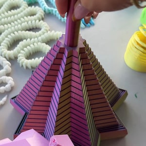 Könnte beinhalten: Eine mehrschichtige, schillernde geometrische Skulptur mit einem zentralen Stab. Die Skulptur wechselt von Rosa zu Gold mit schwarzen Linien. Andere farbenfrohe, abstrakte Formen befinden sich im Hintergrund.