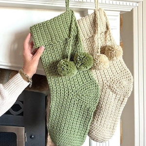 Puede incluir: Dos calcetines de Navidad de crochet con pompones colgados de una repisa de chimenea blanca. El calcetín de la izquierda es verde y el calcetín de la derecha es beige.