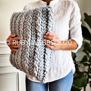 May include: A gray crocheted pillow with a textured pattern. The pillow is being held by a person wearing a white shirt and blue jeans.
