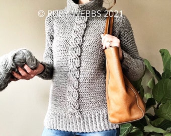 May include: A grey cable knit jumper with a high neck and long sleeves. The jumper is being worn by a person holding a brown leather tote bag. The text "RUBY WEBBS 2021" is visible in the top left corner of the image.