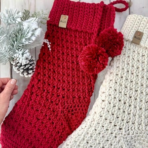 May include: Two crocheted Christmas stockings, one red with a white pom-pom and one white. Both stockings have a brown leather tag with the text "Knot Just Yarn" on them.