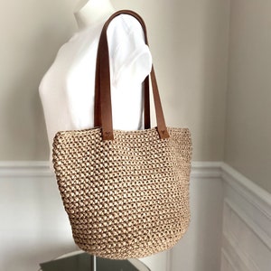 May include: A beige crocheted tote bag with brown leather straps. The bag is large and roomy, ideal for carrying everyday essentials.