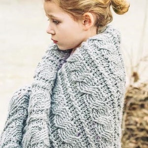 Puede incluir: Una persona joven con una manta de punto gris con un patrón de punto de cable. La manta está envuelta alrededor de los hombros y la cabeza de la persona.