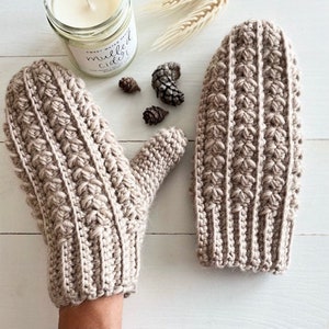 May include: Beige knitted mittens with a textured design. The mittens are shown on a white surface, alongside a lit candle in a jar, pine cones, and wheat. The mittens are a cozy winter accessory.