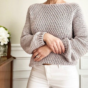May include: A light purple crocheted sweater with a loose fit and long sleeves. The sweater has a textured stitch pattern and a slightly flared hem.