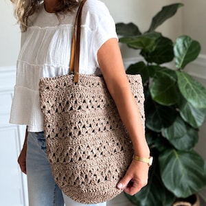 May include: A large, beige, crocheted tote bag featuring an open-work, textured pattern throughout the body. The bag includes two smooth, tan-colored leather straps that are securely attached with metal rivets to the top edge of the bag.