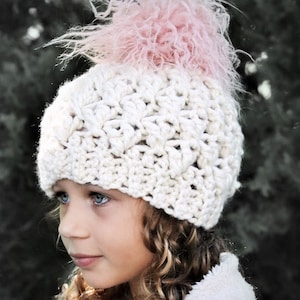 May include: A white crocheted hat with a pink fluffy pom pom. The hat is worn by a child with curly blonde hair.