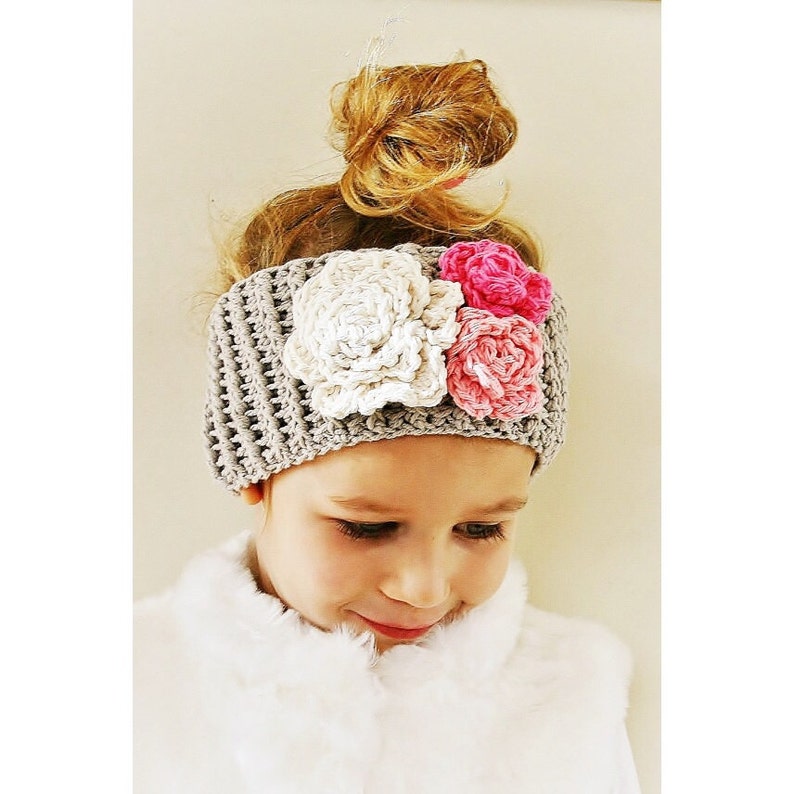 May include: A gray crocheted headband adorned with three large, handmade flowers in cream, pink, and fuchsia. The headband is worn by a young person with light brown hair styled in a bun.