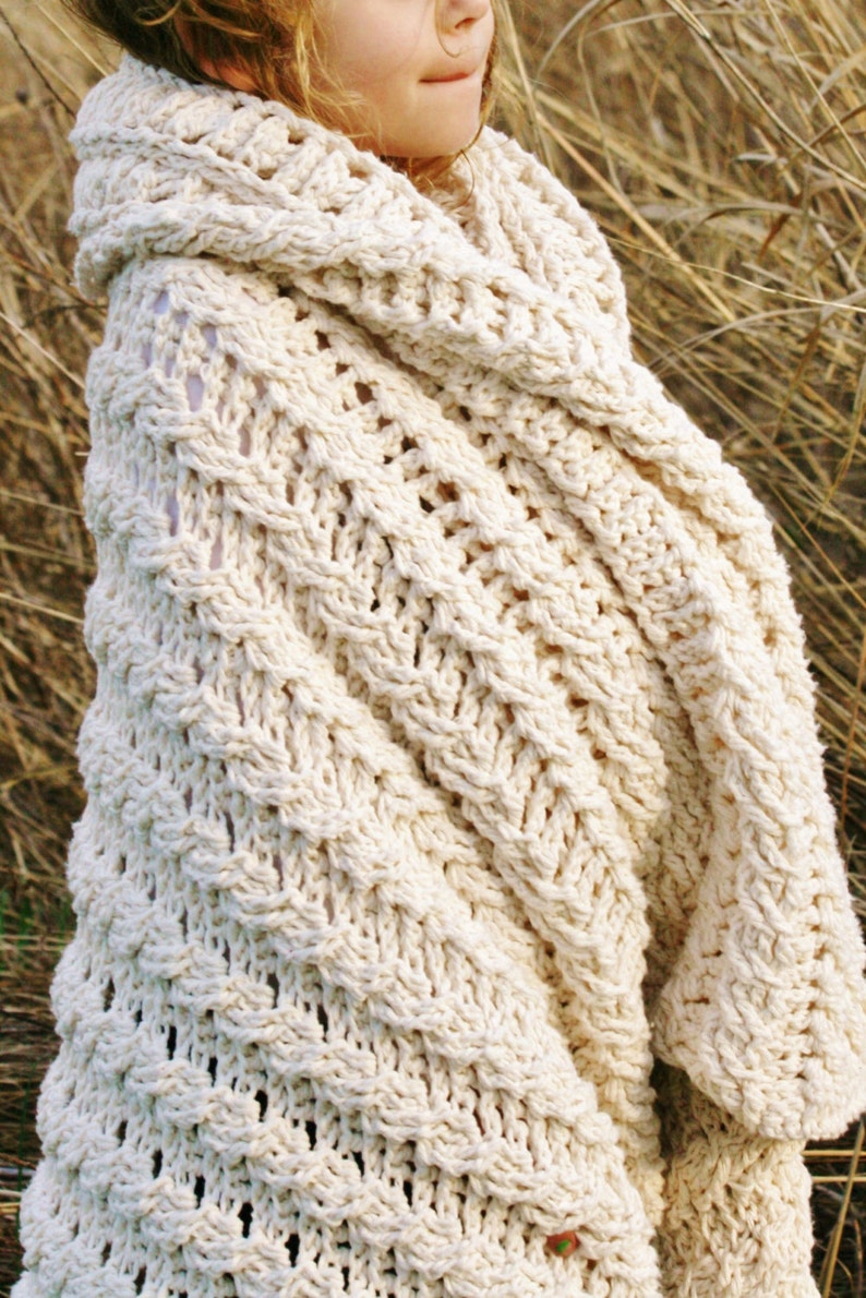 May include: A close-up of a cream-colored crocheted blanket draped over a person's shoulders. The blanket has a textured, open-weave pattern.