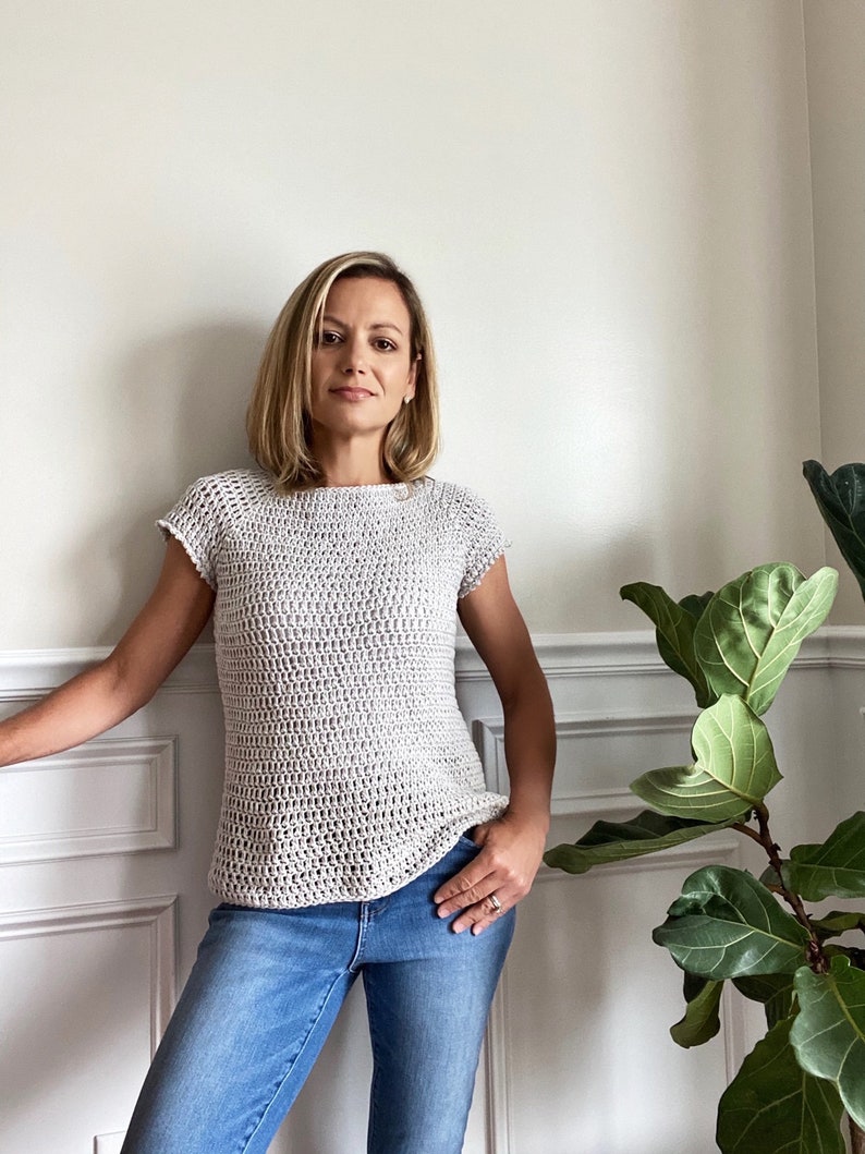 Op de afbeelding: Een vrouw draagt een lichtgrijs gehaakt topje met korte mouwen en een blauwe jeans.