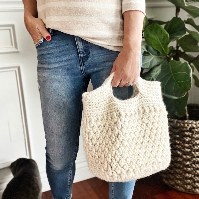 May include: A white crocheted tote bag with a thick, textured design. The bag has a single handle and is being held by a person wearing jeans.
