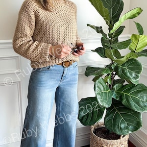 May include: A woman wearing a tan crochet sweater and light blue jeans. She is holding glasses and standing next to a large potted plant with green leaves in a woven basket. The outfit is casual and comfortable.