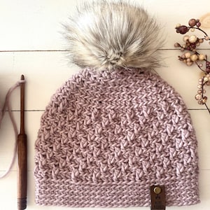 Könnte beinhalten: Eine mauvefarbene Häkelmütze mit einem Kunstfellbommel. Die Mütze hat ein braunes Leder-Etikett mit dem Text "My Cozy Knits" darauf.