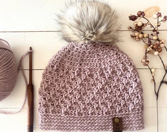 May include: A mauve crocheted beanie with a faux fur pom pom. The beanie has a brown leather tag with the text "My Cozy Knits" on it.