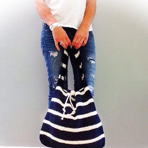 May include: A large, striped crochet tote bag with a white and navy blue color scheme. The bag has a drawstring closure and is being held by a person wearing jeans and a white t-shirt.