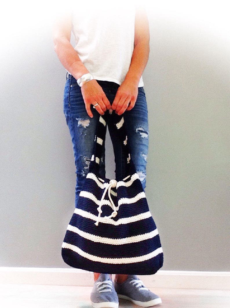 May include: A large, striped crochet tote bag with a white and navy blue color scheme. The bag has a drawstring closure and is being held by a person wearing jeans and a white t-shirt.