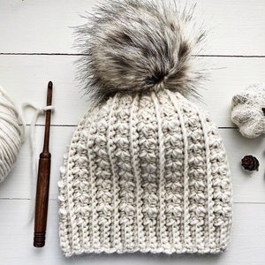 Könnte beinhalten: Eine weiße Häkelmütze mit einem flauschigen braunen Bommel. Die Mütze ist mit einem strukturierten Stich gearbeitet und hat ein schlichtes, klassisches Design.