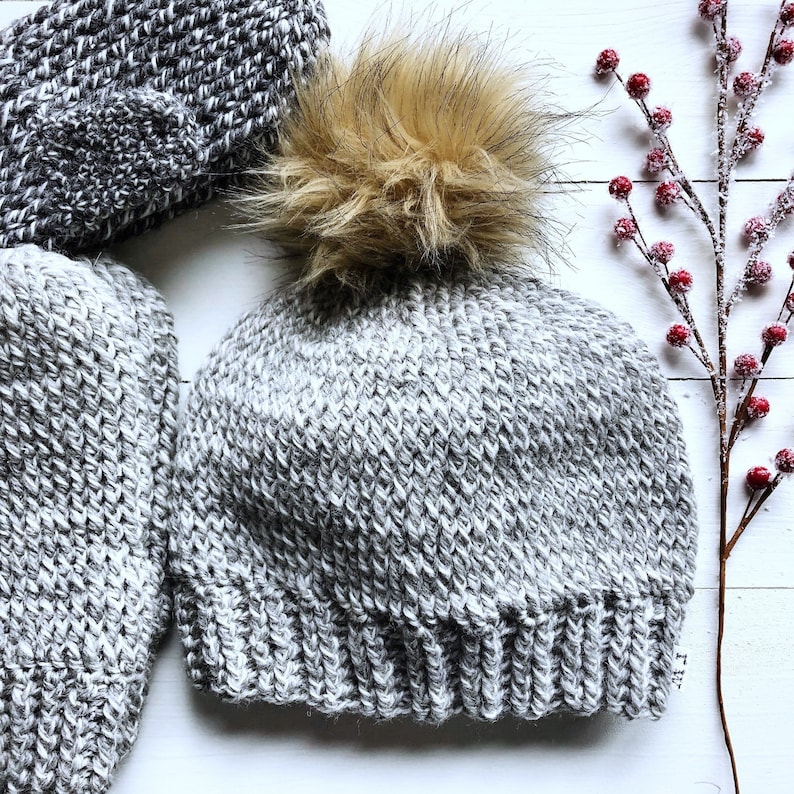 May include: A gray knitted beanie with a fluffy brown pom pom. The beanie is made of a soft, textured yarn.
