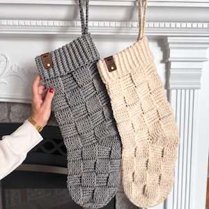 May include: Two hand-knit Christmas stockings, one in charcoal gray and one in cream, featuring a basketweave texture. Each stocking has a ribbed cuff with a small brown leather tag labeled "ruby webbs" and a matching hanging loop at the top.