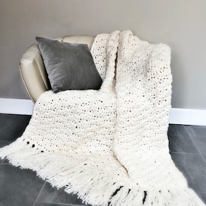 May include: A white crocheted throw blanket with fringe, draped over a cream-colored armchair with a gray pillow.