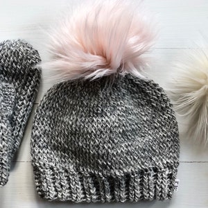 May include: A gray knitted beanie with a pink faux fur pom pom and matching gray knitted mittens.