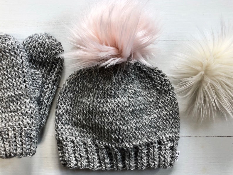 May include: A gray knitted beanie with a pink faux fur pom pom and matching gray knitted mittens.