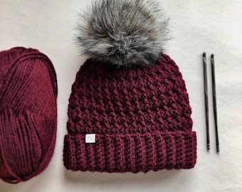 May include: A burgundy crocheted beanie with a faux fur pom pom. The beanie has a white label with the letters "FW" on it. There is a ball of burgundy yarn and two crochet hooks in the background.