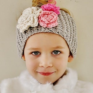 May include: A gray crocheted beanie adorned with three large, handmade flowers in cream and pink. The beanie is worn by a young person with fair skin and blue eyes, wearing a white faux fur vest.