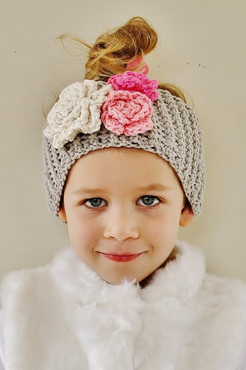 May include: A gray crocheted beanie adorned with three large, handmade flowers in cream and pink. The beanie is worn by a young person with fair skin and blue eyes, wearing a white faux fur vest.