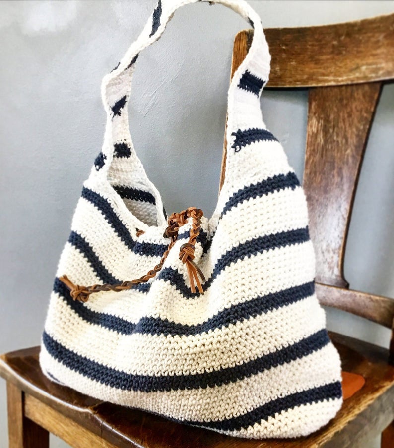 May include: A white and navy blue striped crocheted tote bag with brown leather straps. The bag is sitting on a wooden chair.
