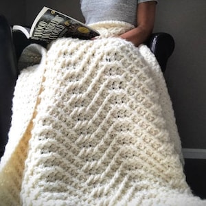 May include: A white crocheted blanket with a chevron pattern. The blanket is draped over a chair and a book is resting on top of it.