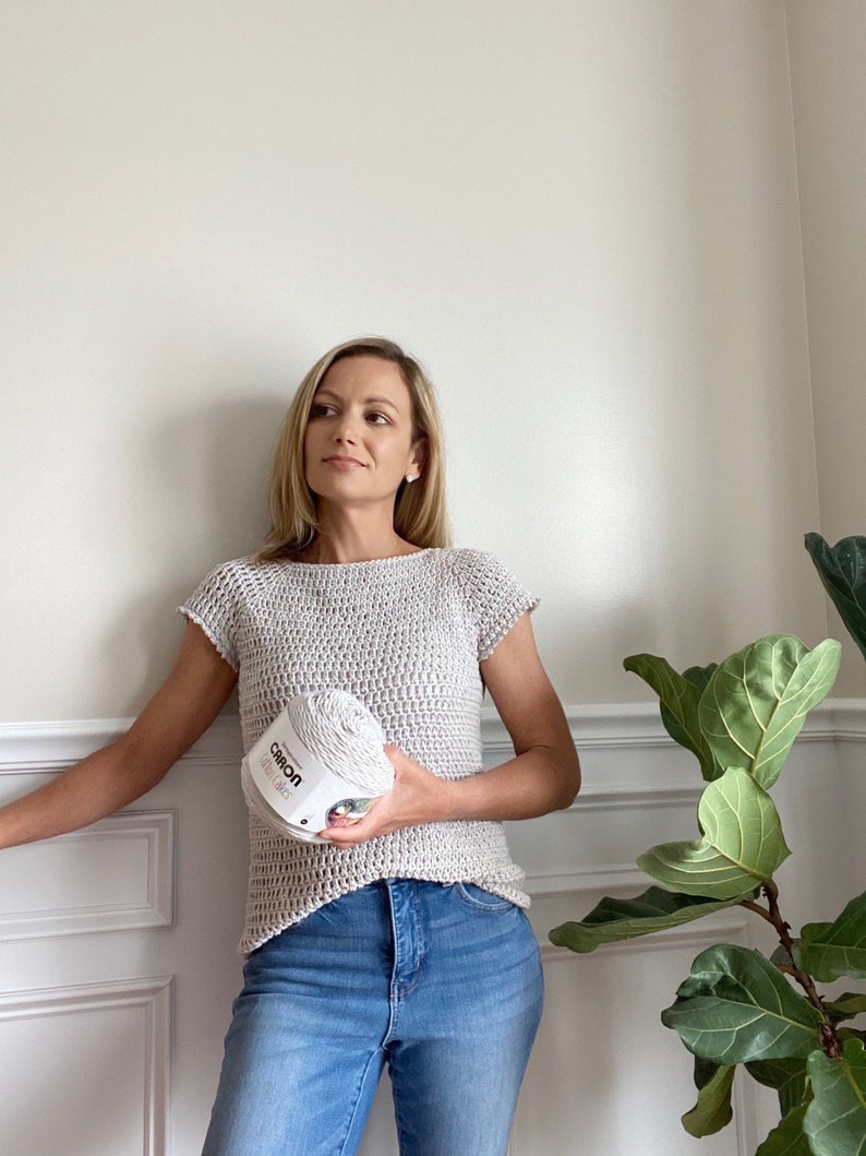 Op de afbeelding: Een vrouw draagt een grijs gehaakt topje met korte mouwen en een blauwe jeans. Ze houdt een bol Caron Simply Soft garen in haar rechterhand.