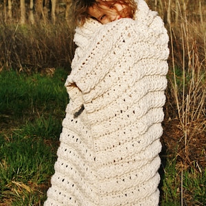 May include: A young person is wrapped in a white crocheted blanket, standing in a field of tall grass.