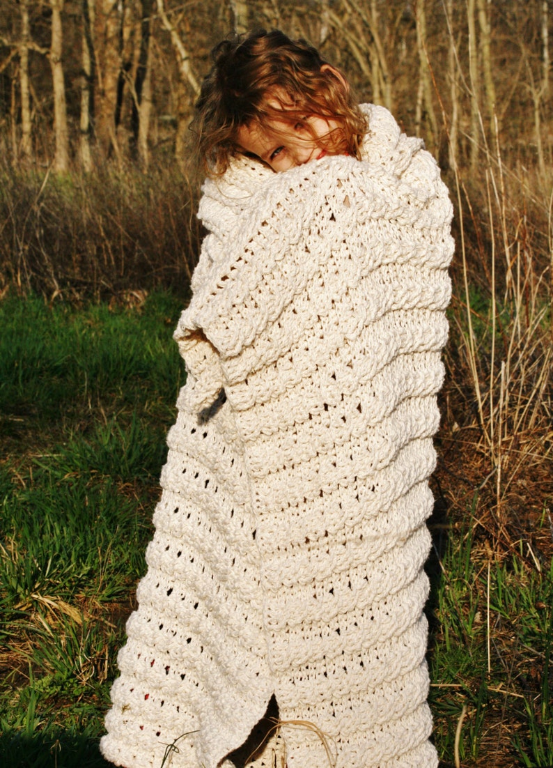 May include: A young person is wrapped in a white crocheted blanket, standing in a field of tall grass.