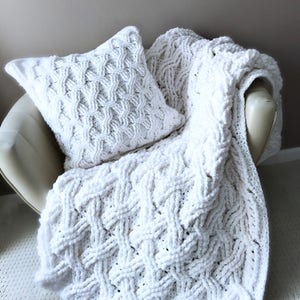 May include: A white, chunky-knit throw blanket and matching cushion rest on a cream-coloured chair. The blanket and cushion feature a textured, woven pattern. The soft, plush blanket is draped over the chair, creating a cosy and inviting aesthetic.