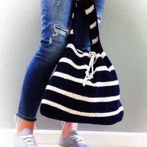 May include: A black and white striped crocheted tote bag with a drawstring closure. The bag is being held by a person wearing blue jeans and gray sneakers.