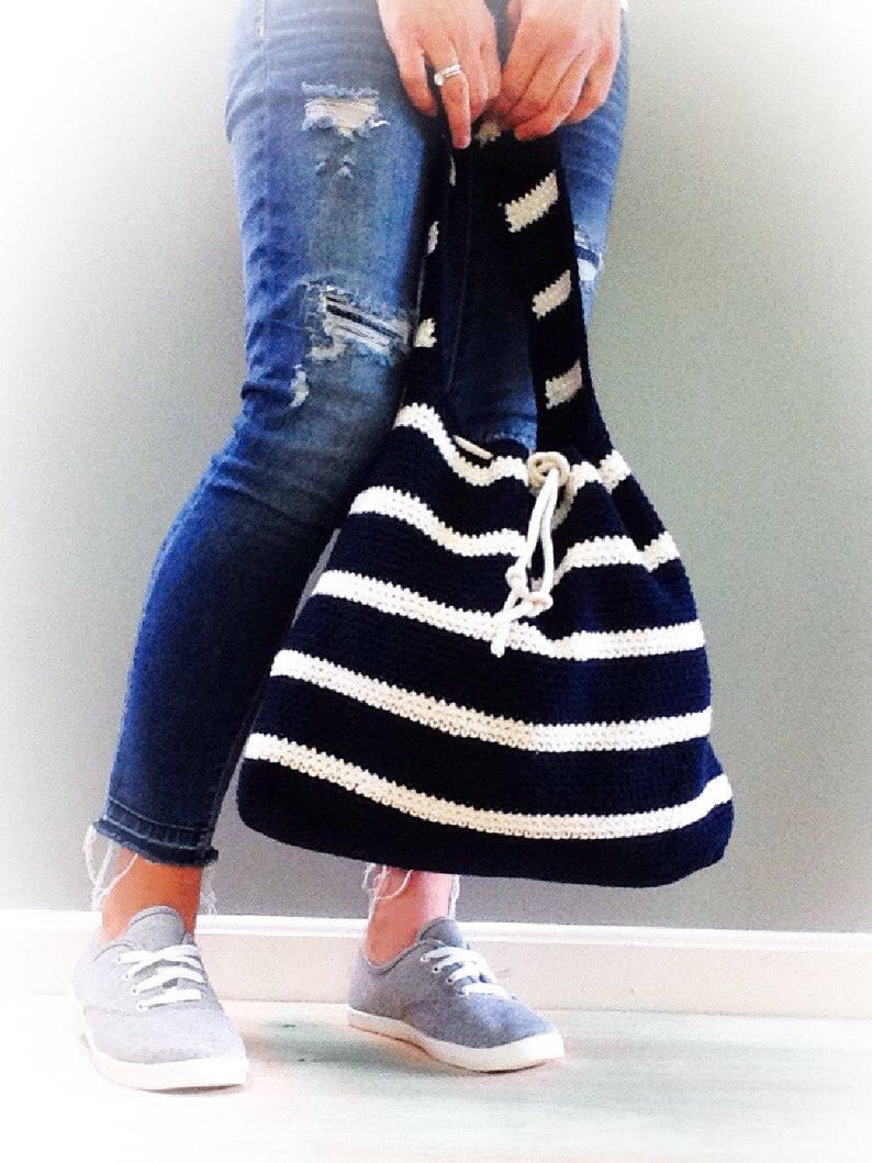May include: A black and white striped crocheted tote bag with a drawstring closure. The bag is being held by a person wearing blue jeans and gray sneakers.