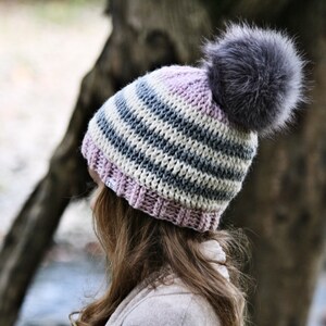 May include: A striped knit beanie with a pink brim and a fluffy gray pom pom. The beanie is made with a combination of white, gray, and light blue yarn.