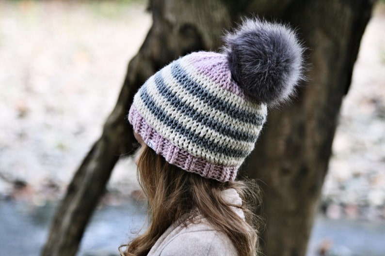 May include: A striped knit beanie with a pink brim and a fluffy gray pom pom. The beanie is made with a combination of white, gray, and light blue yarn.