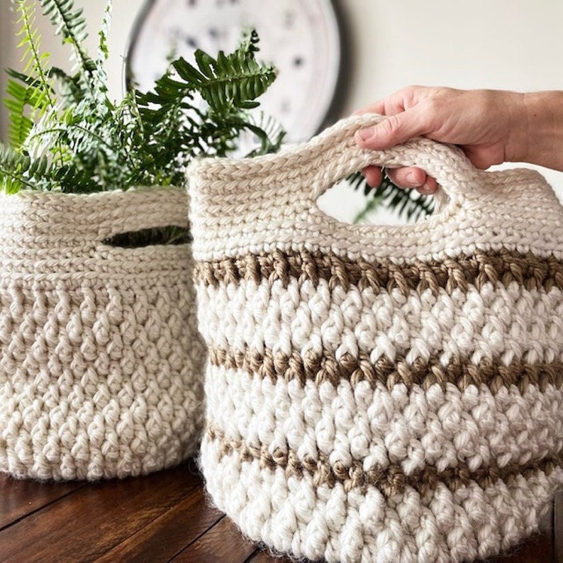 May include: Two crocheted baskets, one with a handle, made with natural fibers. The basket with the handle has brown stripes. The baskets are on a wooden surface.