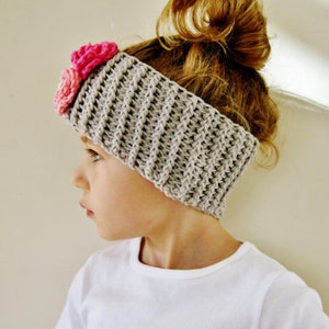 May include: A gray crocheted headband with a textured pattern and two pink fabric flowers. The headband is worn on the head of a person with light brown hair. The headband is a stylish accessory.