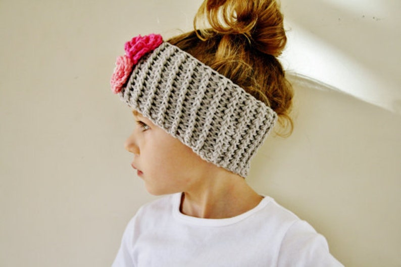 May include: A gray crocheted headband with a textured pattern and two pink fabric flowers. The headband is worn on the head of a person with light brown hair. The headband is a stylish accessory.