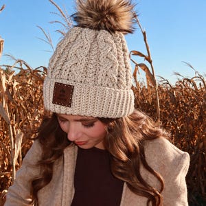 May include: A cream-colored cable knit beanie with a large brown faux fur pom-pom on top. The hat features a ribbed cuff with a square brown leather patch embossed with the text "RUSTY WRENS" and a small tree graphic.