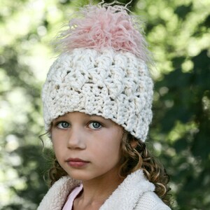 May include: A young girl wearing a white crocheted hat with a pink fluffy pom pom. The hat has a textured design and is worn over her curly hair. She is wearing a white fuzzy jacket.