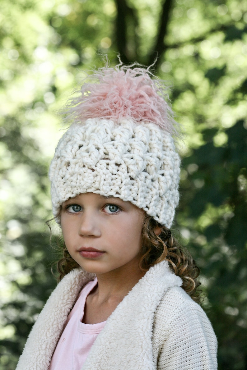 May include: A young girl wearing a white crocheted hat with a pink fluffy pom pom. The hat has a textured design and is worn over her curly hair. She is wearing a white fuzzy jacket.