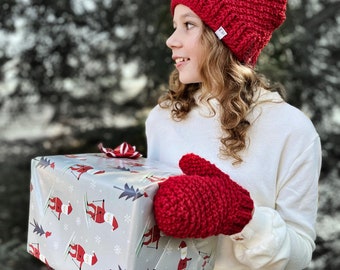 May include: A young person wearing a red knit hat with a faux fur pom pom and red knit mittens holds a wrapped gift with a Santa Claus pattern. The gift is wrapped in gray paper with white snowflakes and red accents.