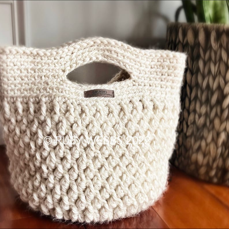 May include: A crocheted cream-colored tote bag with a brown leather handle and a brown leather label that says "Ruby Webb's 2022".